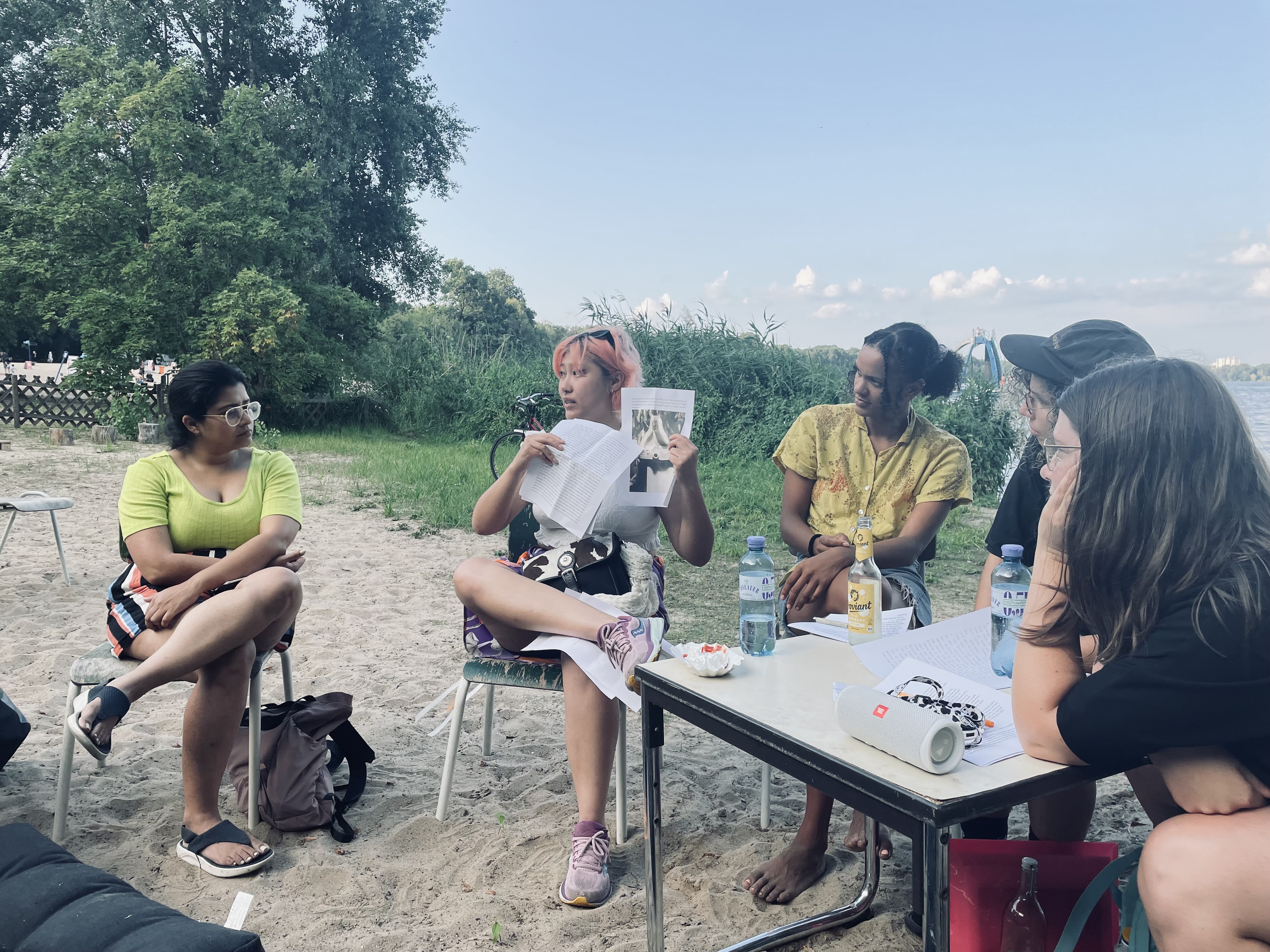 Image of Alungo Xatan, a brown-skinned person with orange hair, talking into a microphone to a circle of listeners.  They are all seated outdoors at a lake next to the water.