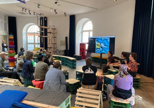 Image of a group sitting down in an art gallery watching three people give a presentation.  There is a cartoon minion on the screen of the presentation.