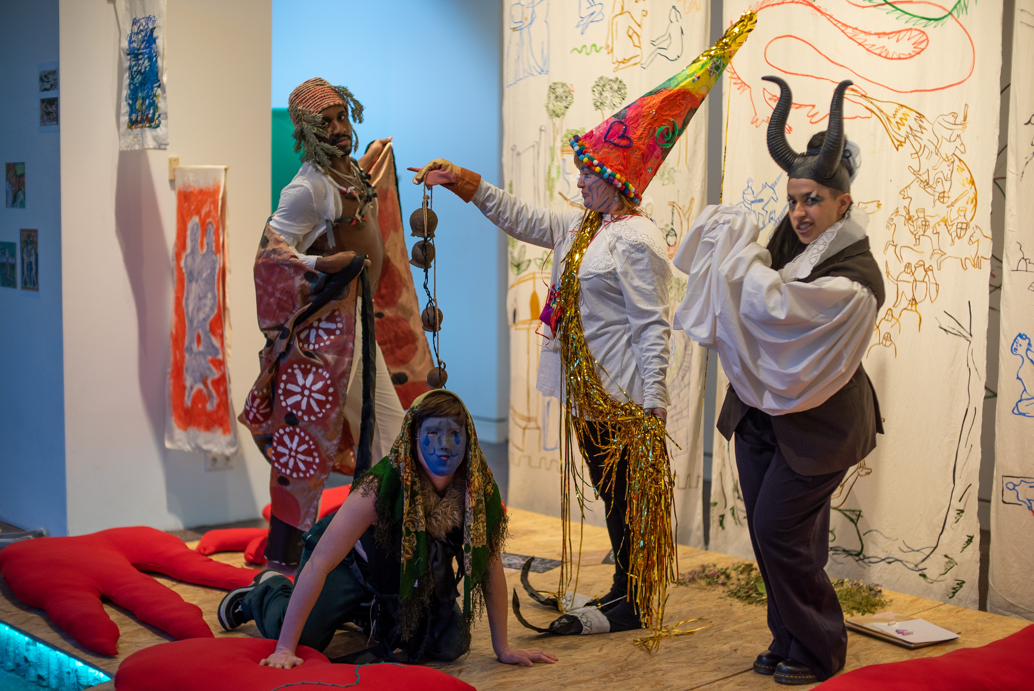 Image of four people on a colourful stage in a gallery. They are wearing oversized princess hats, devils horns, handpainted capes and masks, and surrounded by hand-sewn red cushions and paintings with medieval themes