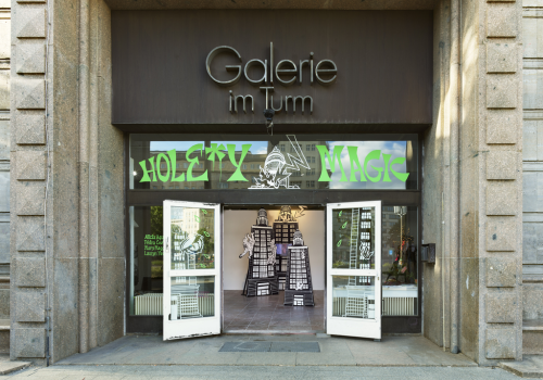 The doors of a gallery with signs reading Galerie im Turm and Hole*y Magic. A sculptural installation can be seen inside.