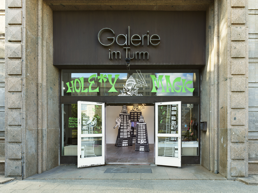 The doors of a gallery with signs reading Galerie im Turm and Hole*y Magic. A sculptural installation can be seen inside.